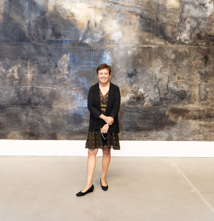 A woman stands smiling in front of a large abstract painting with dark, muted tones of gray, brown, and black. She is wearing a patterned dress with a black cardigan and black flats, holding a face mask in her hands. The floor is a smooth, light gray concrete.