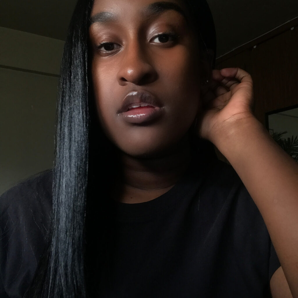 Close-up of a person with smooth, straight black hair falling over one shoulder, wearing a black shirt. Their lips are slightly parted and glossy, and their hand is raised to touch the side of their head. The background is dimly lit with a wooden panel and a mirror partially visible.