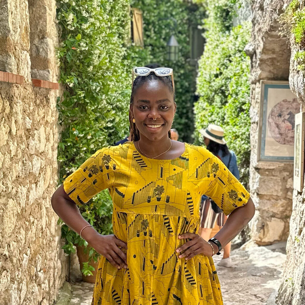 A smiling woman stands in a narrow stone alleyway with lush green foliage on both sides. She wears a bright yellow dress with a geometric and plant pattern, sunglasses resting on her head, and has her hands on her hips. In the background, a person wearing a hat and striped shorts is partially visible walking away. The setting appears sunny and vibrant.