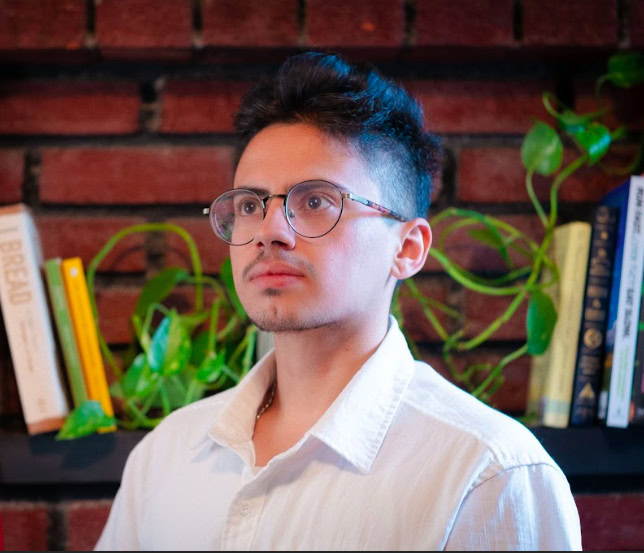 A young man with short, dark hair and a trimmed beard is wearing round glasses and a white collared shirt. He is looking slightly to the side with a thoughtful expression. Behind him is a brick wall with a shelf holding several books and green leafy plants.