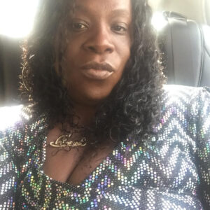 A person with long curly hair is sitting in a vehicle, wearing a sparkling top with a zigzag pattern. They have a neutral expression and theres a seatbelt visible beside them. The photo is taken from a close angle.