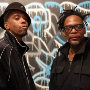 Two people stand in front of a wall with abstract spray-painted graffiti. Both wear black jackets; one has a black hat and the other has black glasses. They appear confident and serious.