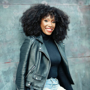Smiling person with curly hair wearing a black leather jacket, black top, and distressed jeans sits against a textured gray background.