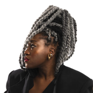 A woman is depicted in profile, showcasing a striking hairstyle with long, twisted gray and black hair styled high. She wears bold, gold hoop earrings and has a deep red lip color. Her outfit features a black blazer with structured shoulders, adding to her sophisticated appearance. The background is plain white, emphasizing her features and style.
