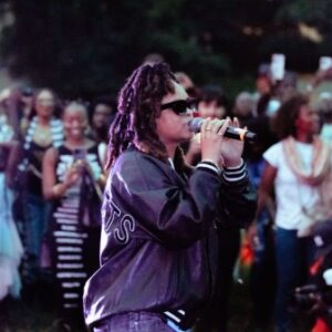 A performer wearing sunglasses and a black jacket sings into a microphone outdoors, surrounded by a large, diverse crowd of people, many of whom are taking photos or videos.