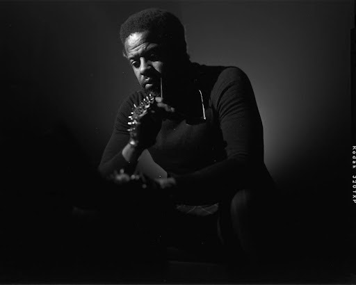 A man in a dark turtleneck sits pensively in low lighting, with one hand resting on his chin and an arm adorned with spiked accessories, creating dramatic shadows across his face and body.