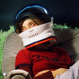 Person wearing a white astronaut helmet with a blue visor, a brown knit hat underneath, a red shirt with black text, and light-colored pants, reclining on a gray cushioned chair with a green feathered trim.