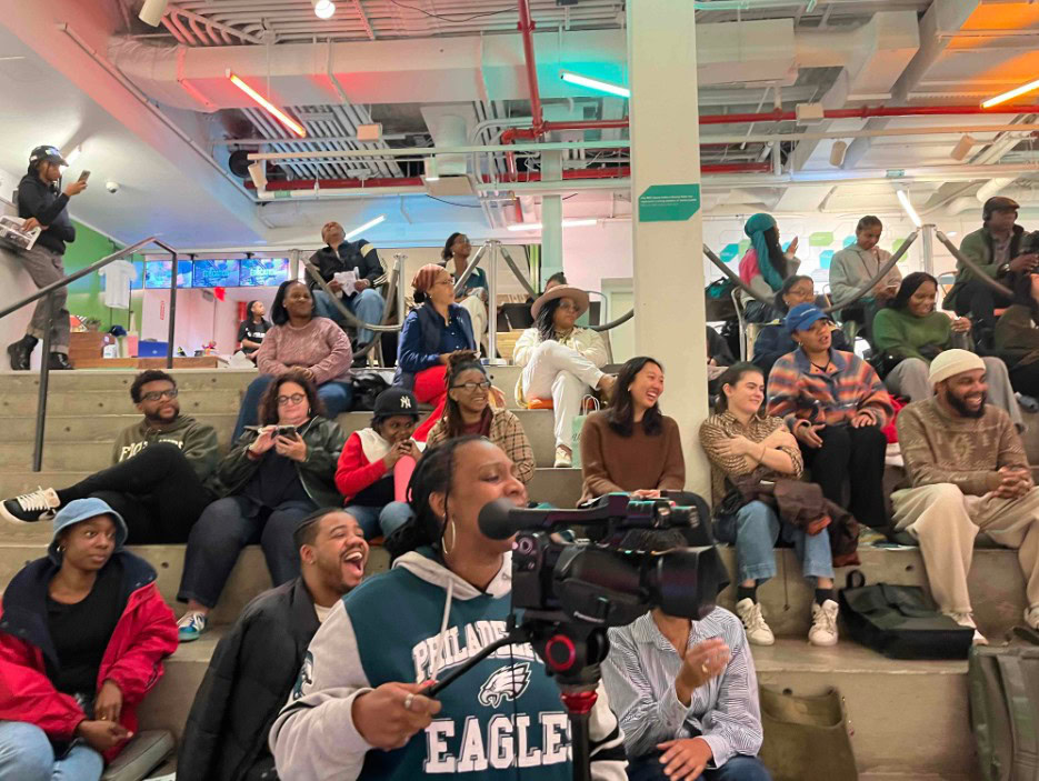 A diverse group of people seated on tiered concrete steps in an indoor space, some smiling and laughing. The foreground features a person wearing a Philadelphia Eagles hoodie operating a video camera on a tripod. The ceiling has exposed pipes and colorful neon lights. Some individuals are engaged with their phones, while others are interacting with each other.