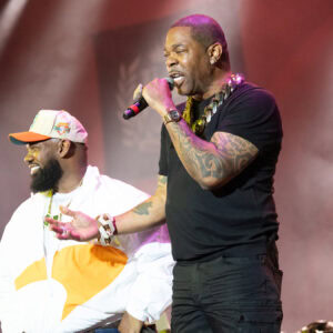 Two male performers on stage, one wearing a white and orange jacket with a white and orange cap, smiling and looking to the side, the other wearing a black t-shirt and black pants, holding a microphone and singing, with tattoos on his arms and a large gold chain necklace. The background is smoky with red lighting.