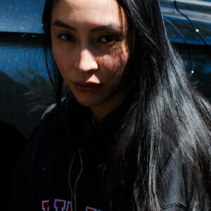A young woman with long black hair and a serious expression stands outside in partial sunlight, wearing a dark hoodie. A black car is visible in the background.