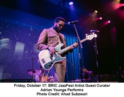 Musician Adrian Younge plays an electric bass guitar on stage under colorful lights at BRIC JazzFest, with a drummer and stage backdrop behind him.