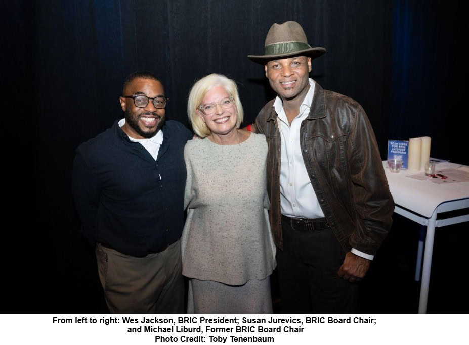 Three people stand together smiling in front of a dark backdrop. On the left is a man wearing glasses, a dark shirt, and light-colored pants. In the center is a woman with short blonde hair and glasses, wearing a light gray speckled top and matching pants. On the right is a man wearing a brown leather jacket, white shirt, dark pants, and a greenish-brown fedora hat. A table with candles and a small sign is visible in the background on the right side.