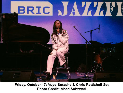 A woman sits on a stool, singing into a microphone on stage beside a piano and drum set. The backdrop reads BRIC Jazzfest. Stage lighting casts a purple-blue hue. Text below credits Vuyo Sotashe & Chris Pattishell, photo by Ahad Subzwari.