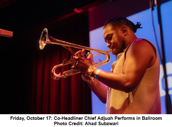 A musician plays a trumpet on stage under purple lighting, wearing a sleeveless top and necklace. The background shows a projected blue and white image.