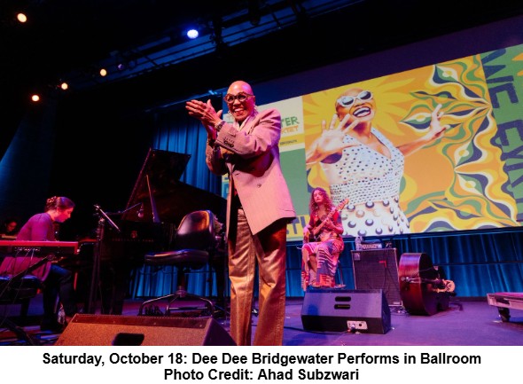 A jazz musician in a suit sings and claps on stage beside a piano, upright bass, and other performers. Behind him is a large, colorful image of a smiling woman. Stage lighting is bright and vivid.