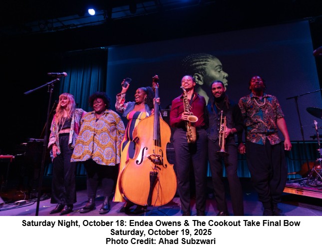 A band of six musicians stands side by side on stage, smiling and holding hands. One member holds an upright bass. A large projected portrait is visible behind them. Stage lights cast a vibrant glow.