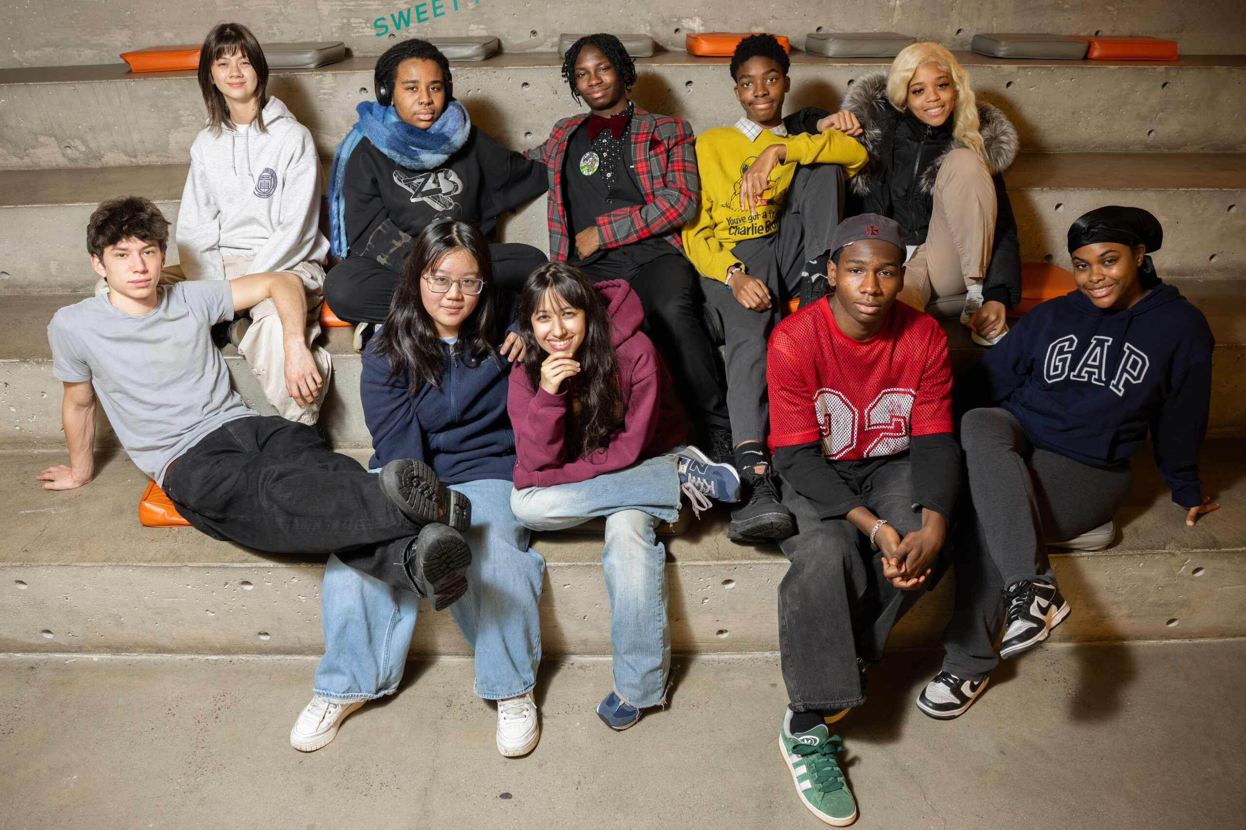 A diverse group of young people sitting on concrete steps. They are dressed casually in a variety of clothing including hoodies, jackets, sweaters, and jeans. Some are smiling, and one person wears headphones and a blue scarf. The background is a plain concrete wall with cushions placed on the steps.