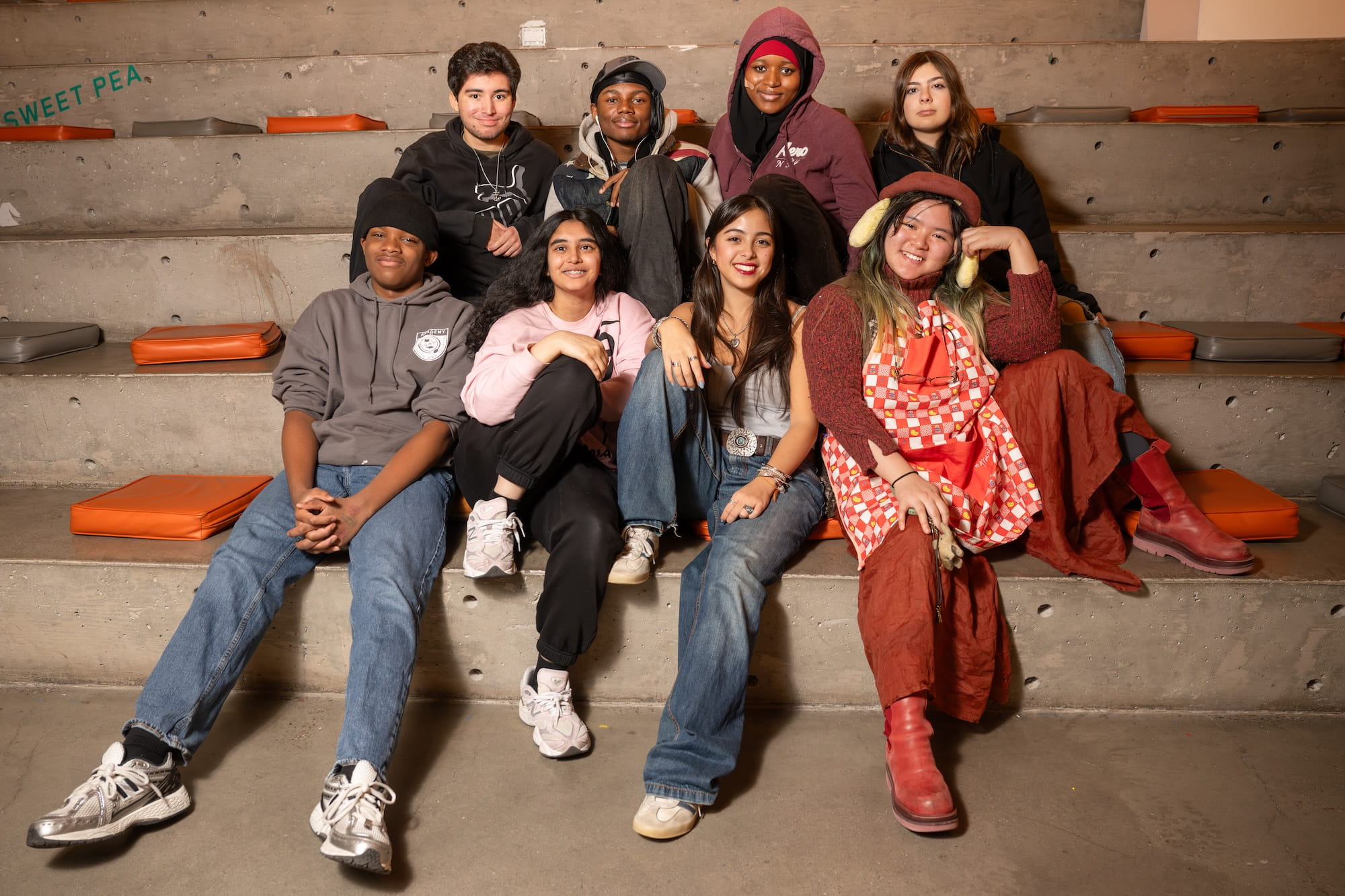 A group of seven young people sit and smile on concrete bleachers with orange cushions. They wear casual, colorful clothing and appear relaxed and happy together.