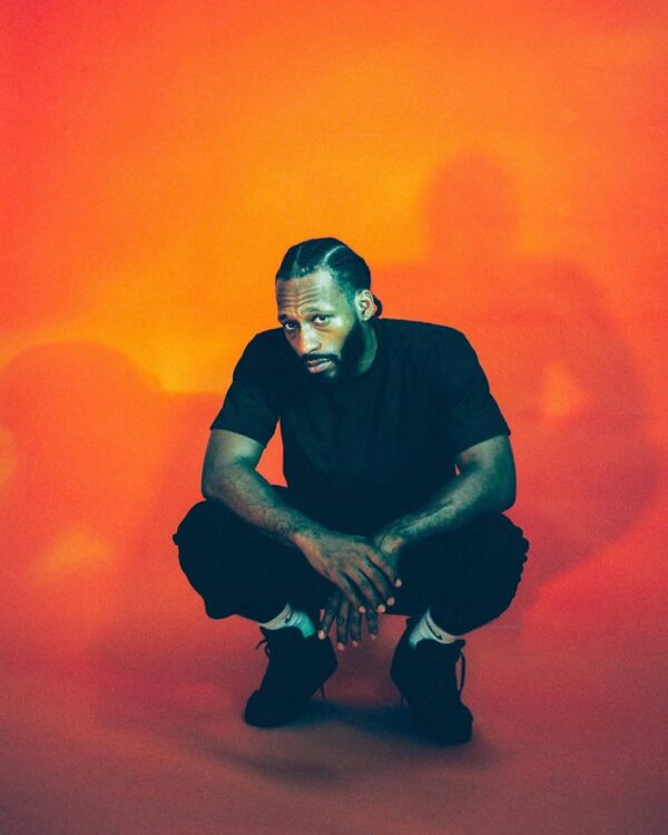 A man with braided hair and a beard is squatting against a vibrant orange background. He is wearing a black t-shirt, black pants, black shoes, and white socks. His hands are clasped together in front of him, and he has a serious expression on his face.