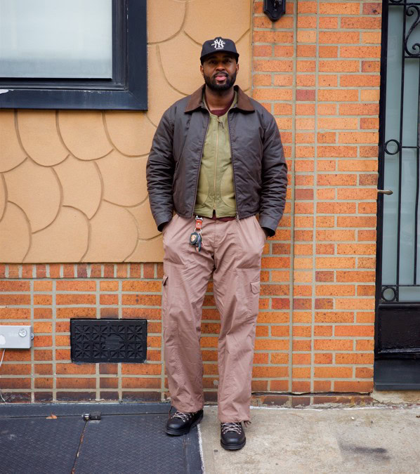 A man stands against a brick and textured wall wearing a black New York Yankees baseball cap, a dark brown jacket over a green zip-up jacket, light brown cargo pants, and black shoes. He has his hands in his pockets and keys hanging from his pants.