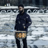 https://bricartsmedia.org/wp-content/uploads/2026/07/Standing-with-Drum-Outside.jpg
